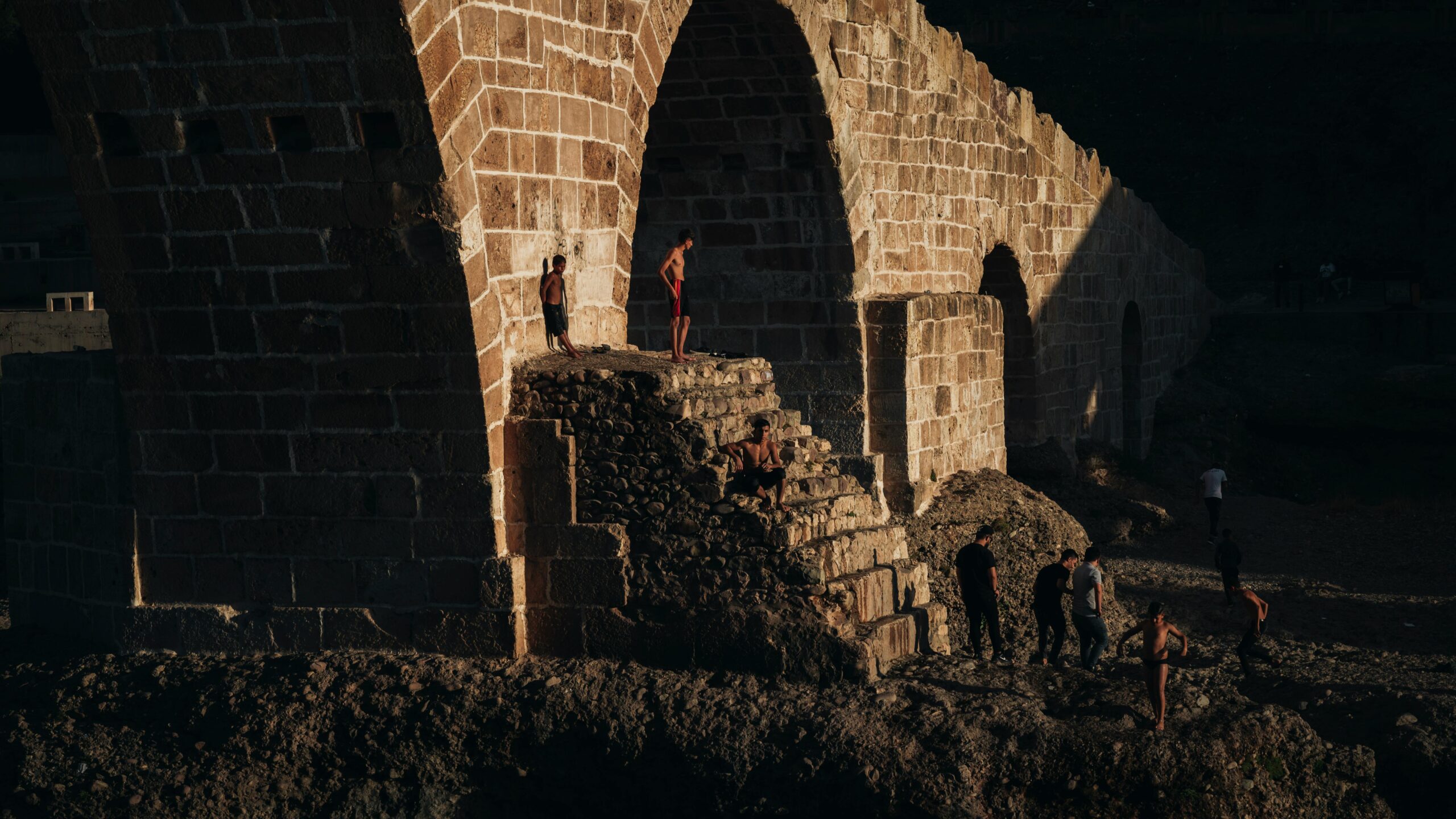 People gather around the historic Pira Delal stone bridge in Zakho, Iraq at sunset.